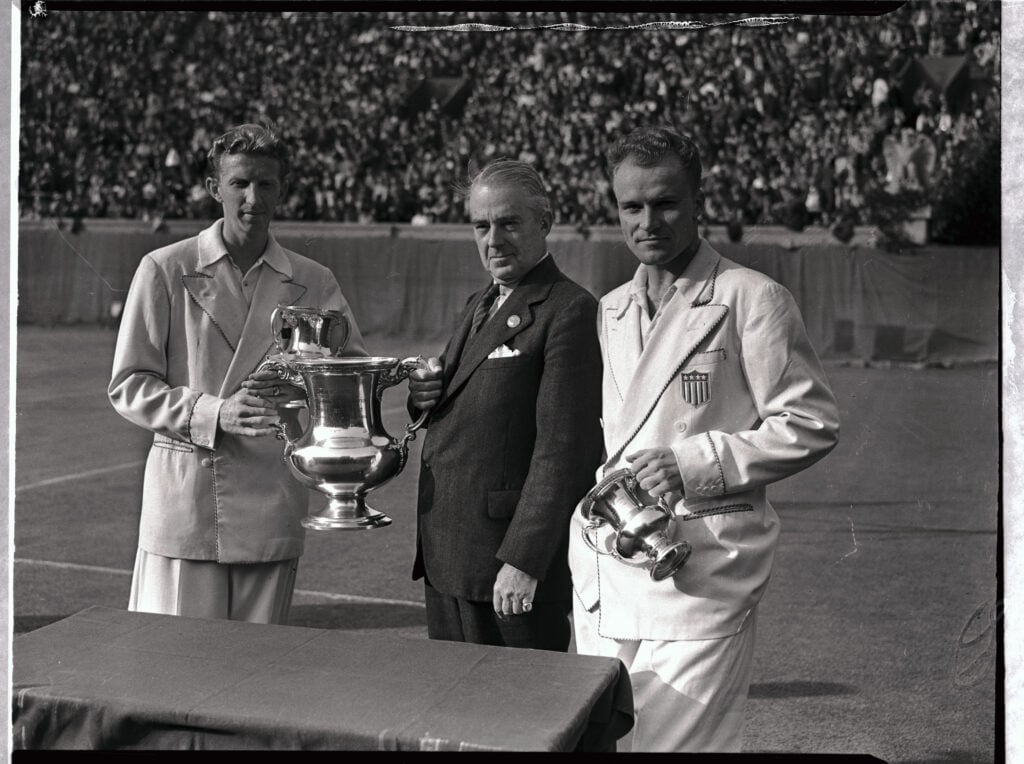Donald Budge, world's number one tennis player, receiving the cup, emblematic of the U.S. Men's Singles Tennis Championship, from Holcombe Ward, president of the U.S. Lawn Tennis Association, watched by Gene Mako, whom he defeated, 6-3, 6-8, 6-2, 6-1, in their finals match today. Budge's victory gave him a clean sweep of the four major titles in international net competition - the British, the French, the Australian, and the American championships.